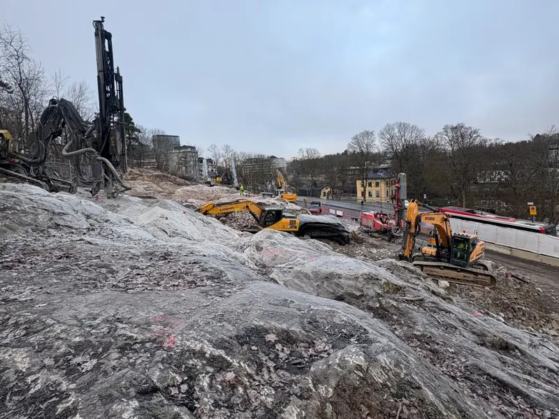 Bergarbete med borrigg och grävmaskiner intill väg och bebyggelse.
